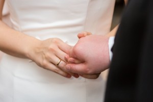Frauenhand mit goldenem Ehering, Ausschnitt von Frau im Brautkleid und Mann im dunklen Anzug. Beide reichen sich die Hände zur Eheschließung.